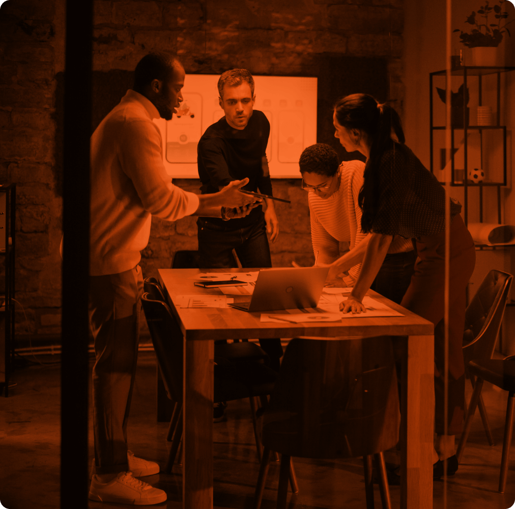 Four people discussing work while standing around a table in a meeting room.